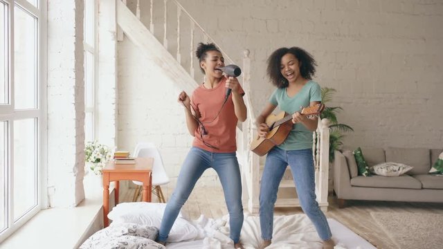 Mixed race young funny girls dance singing with hairdryer and playing acoustic guitar on a bed. Sisters having fun leisure in bedroom at home