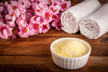 Wellness setting. Sea salt in bowl, towel and flowers on brown textured background. Selective focus. Place for text. Flat lay.