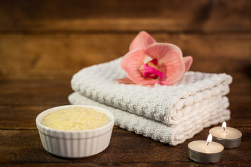Obraz na płótnie Canvas Spa or wellness set. Yellow sea salt in white bowl, towels, candle and pink flowers lily on brown wooden background. Selective focus. Place for text. Health concept.