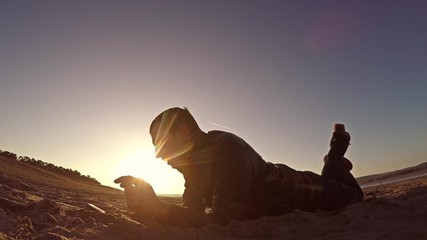 Portable console. Man lies on the sand and plays in the portable console at sunset sunlight