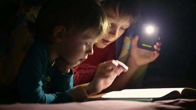 Children read a book under a blanket with a flashlight