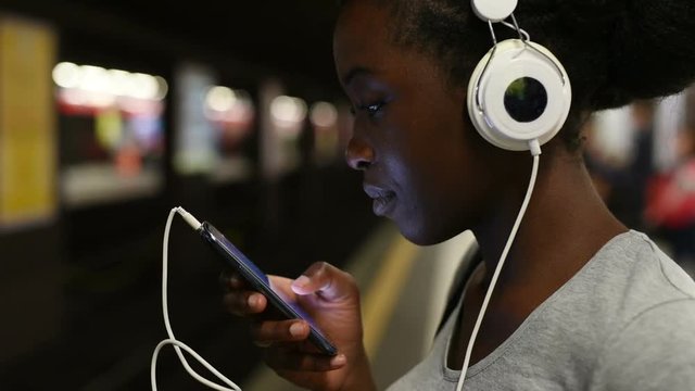 Half length of young beautiful afro black woman in underground, listening music with headphones and tapping screen of smart phone hand hold - technology, relaxing, social network concept