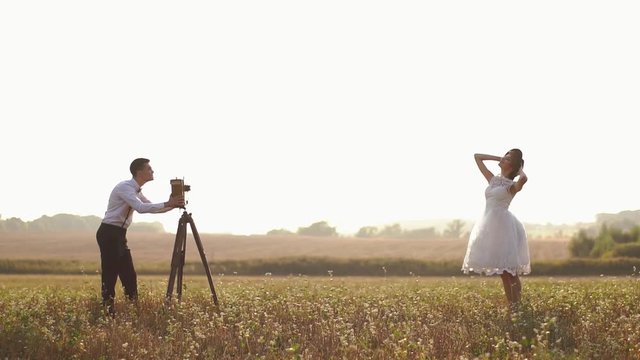 The side view of the groom taking pictures of the posing bride using the old vintage camera in the sunny field.