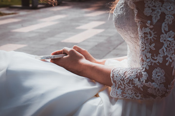 Tender bride in white dress with lace is holding phone in her hands. Beauty model girl in wedding day. Hands close-up in the park