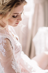 Fototapeta na wymiar Bride's morning. Fine art wedding. Portrait of a young bride in white lace boudoir with wavy blonde hair and a bouquet in her hands posing looking down shyly