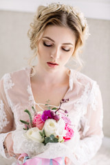 Bride's morning. Fine art wedding. Portrait of a young bride in white lace boudoir with wavy blonde hair and a bouquet in her hands posing looking down shyly