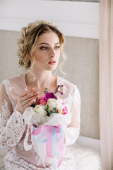 Bride's morning. Fine art wedding. Portrait of a young bride in white lace boudoir with wavy blonde hair and a bouquet in her hands posing looking down shyly