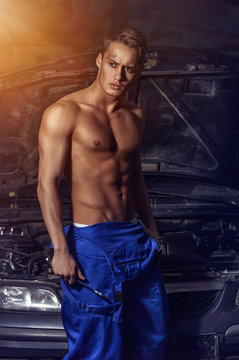 A mechanic man with a muscular naked torso stands near a car with an open hood in the bright light of a searchlight
