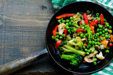 Frying pan with fried egg and a vegetable mixture: peanuts, broccoli, paprika and frozen broccoli on the side on natural wooden background, top view