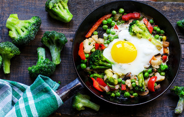 Frying pan with fried egg and a vegetable mixture: peanuts, broccoli, paprika and frozen broccoli on the side on natural wooden background, top view