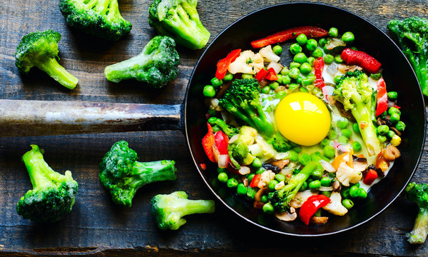 Frying pan with fried egg and a vegetable mixture: peanuts, broccoli, paprika and frozen broccoli on the side on natural wooden background, top view