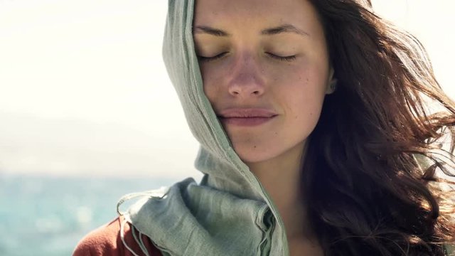 young woman with scarf on head and closed eyes standing in wind against a sea landscape