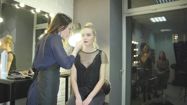 Young woman makeup artist removes excess cosmetic from the face blonde client at the beauty salon next to a large mirror before a white brick wall.
