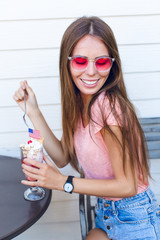 Close-up of cute girl sitting on a chair with white background behind eating ice-cream with cherry on top with a spoon. She wears denim shorts, pink top and smiles. She has pink eyeglasses.