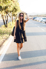 Obraz premium Stylish girl standing near road wearing short black dress, straw hat, black eyeglasses, white sneakers and black backpack. She smiles and holds her hat with hand.