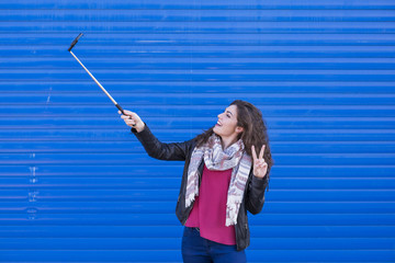 Happy beautiful woman taking a selfie with smart phone over blue background. Wearing casual clothing. Selfie stick