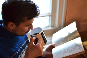 Young handsome man in a knitted sweater reading a book with a Cup of coffee, tea in hand at the window, cozy atmosphere, pleasant surroundings, self-education