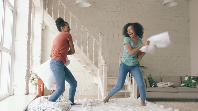 Slow motion of two mixed race young pretty girls jumping on bed and fight pillows having fun at home