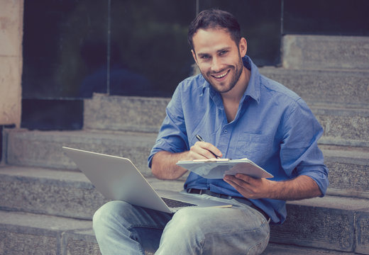 A handsome man sitting on steps with laptop and a notepad