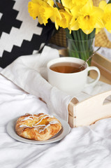 Wooden tray with light breakfast and flowers on white textile in bed. Herbal tea Breakfast in bed Morning Yellow daffodils.