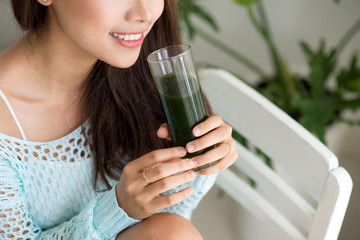 Pretty young asian woman holding green fresh vegetable juice or smoothie from glass at home