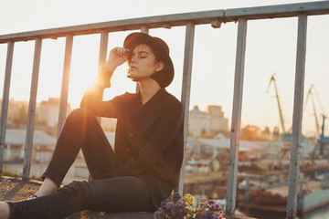Waiting on the bridge. Black style teen girl sitting on the ground. Urban evening port on...