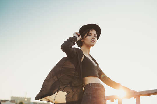 Adorable young woman in stylish black clothes and hat looking to the cam. Evening sunset atmosphere of the city