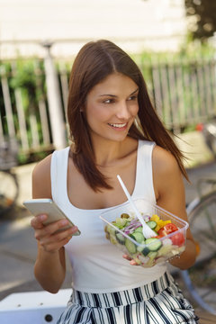 frau isst einen salat to-go und hält ihr handy in der hand