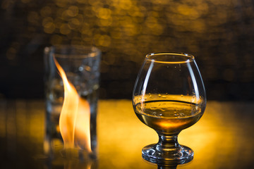 Glass o whisky with ice and wineglass of coghac and fire flames on yellow bokeh background