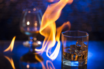 Glass o whisky with ice and wineglass of coghac and fire flames on bokeh background
