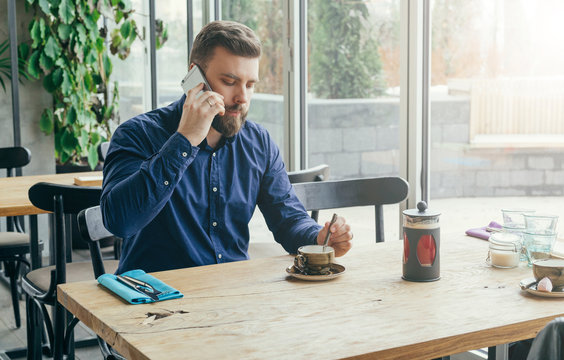 Young bearded businessman in blue shirt is sitting at wooden table near window in restaurant and talking on cell phone.
