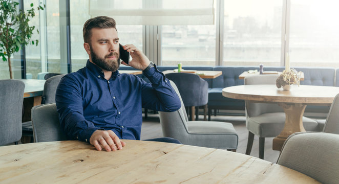 Bearded attractive businessman in blue shirt is sitting at wooden round table in restaurant and talking on cell phone.