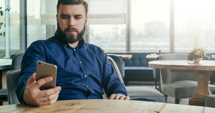 Young bearded businessman in blue shirt is sitting at table in restaurant and is using smartphone.