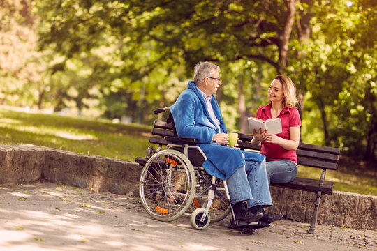 Young carer woman reading book outdoor disabled man in wheelchair.