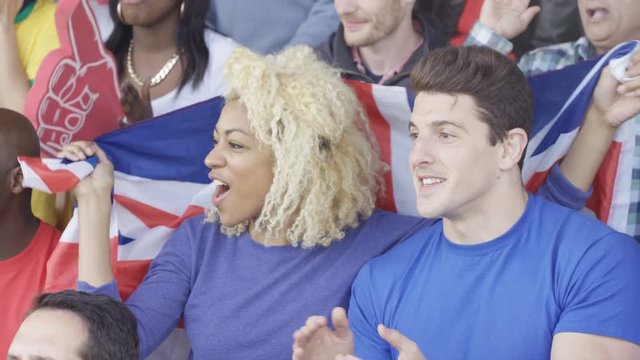  Couple sitting in the crowd at sports event, young woman holding British flag