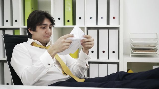 Businessman sitting at a table, reading a report and making a paper plane from it. Concept of futility of life. Locked down real time medium shot