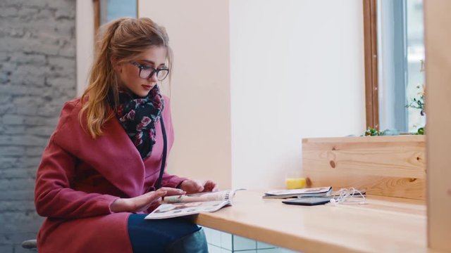 Cozy caf&eacute;&rsquo;s interior, attractive European girl in a stylish casual wear sitting by the window and flipping the pages of the magazine. Modern lifestyle, active breakfast.