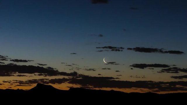 Static timelapse of a sickle moon setting at dusk with scattered clouds and a silhouetted mountain range available on request.