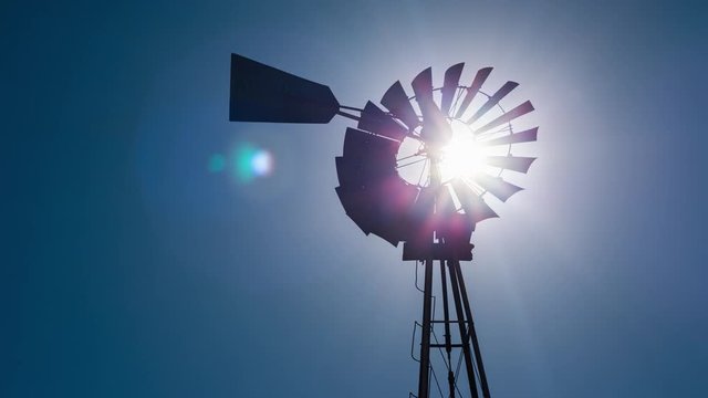 Static daytime timelapse of a silhouette Windmill frantically blowing in the wind against a blue sky and the bright sun