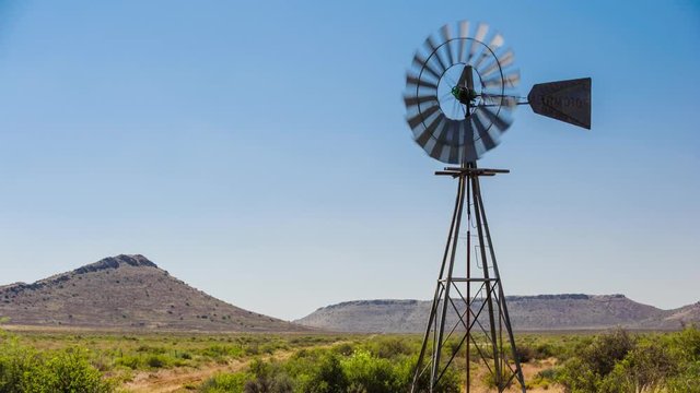 A static timelapse of a typical Karoo landscape scene filled with shrubs and grass with a windmill blowing frantically in the wind against a bright blue sky and hills in the background available on request.