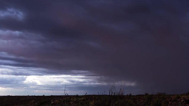 A static timelapse of an intense thunderstorm with dark and dramatic clouds and lightning flashes as the rain is pouring down the landscape with a few rocks and shrubs in the foreground