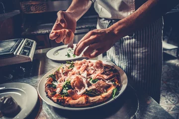 Schilderijen op glas Food concept. Preparing traditional italian pizza. Chef in uniform decorate ready dish with green rucola herbs, ham, salami and prosciutto in interior of modern restaurant kitchen. Ready to eat. © Goinyk