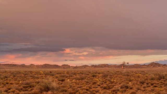 A beautiful static timelapse of a typical Karoo farm landscape with a windmill in soft warm magenta light with a various cloud movements, typical of an old artist painting
