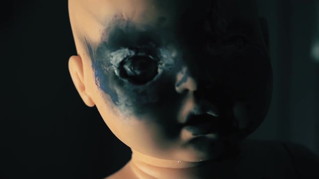 A wicked doll, moving its burnt eyes and mouth, looking at the viewer and following him until everything is dark. Handheld travelling shot.  Halloween horror themed clip.
