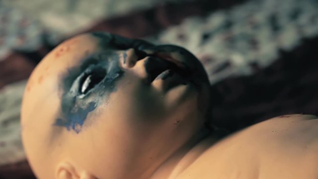 A wicked doll, moving its burnt eyes and mouth, lying on a bed. Handheld travelling shot.  Halloween horror themed clip.
