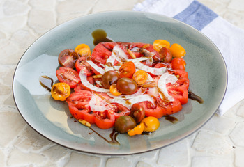 A vibrant healthy mixed tomato, mozzarella, pine nut and balsamic vinegar salad, served on a rustic plate on a tile table.