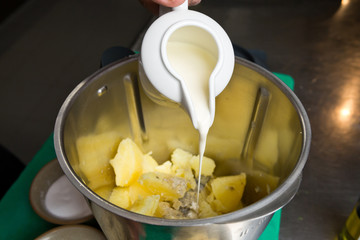 Close up shot of a jug of cream, being poured onto fluffy peeled potatoes in a blender.