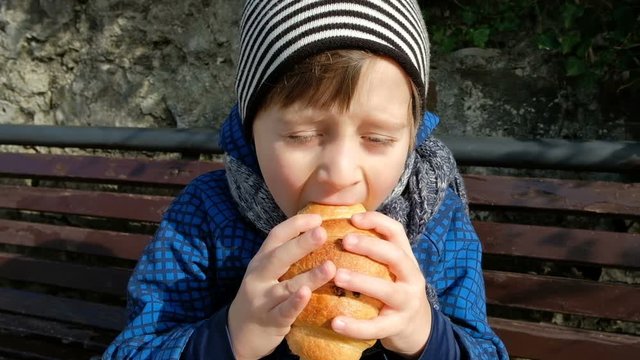 Little boy enjoy eating large croissant sitting on bench outdoor. Caucasian child in blue plaid jacket with hood, knitted scarf, white cap with black stripes grab with both hands fresh baked good