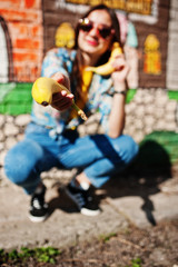 Fototapeta premium Beautiful fun teenage girl speaking bananas like phone, wear yellow t-shirt, jeans and sunglasses near graffiti wall.