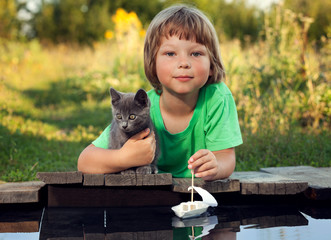 boy and his beloved kitten playing with a boat from pier in pond summer evening